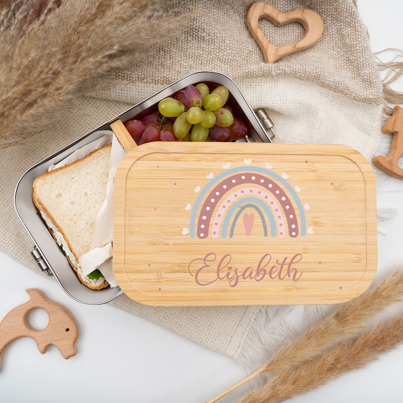 Może przedstawiać: Drewniana lunchbox z wzorem tęczy i imieniem "Elizabeth" na pokrywce. Lunchbox jest otwarta, a wewnątrz widać kanapkę i winogrona.
