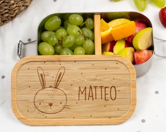 Peut inclure: Une boîte à lunch en bois avec un intérieur en acier inoxydable. Le couvercle présente un joli motif de lapin et le nom "MATTEO" gravé dessus. La boîte à lunch est remplie de fruits, notamment des raisins, des pommes et des oranges.
