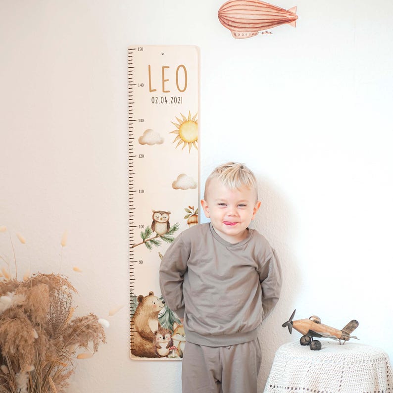 May include: A wooden height chart featuring a printed scale from 50 to 150 cm, the name LEO, and the date 02.04.2021. The design includes illustrations of a sun, clouds, an owl, a bear, a hedgehog, and a fox in soft, muted colors.