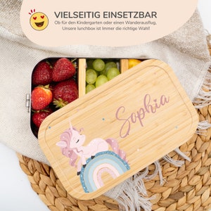 May include: A pink and white unicorn with a rainbow tail sits on a wooden lunchbox with the name "Sophia" written on it. The lunchbox is made of bamboo and has a stainless steel interior.