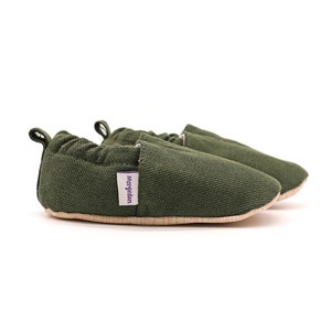 Puede incluir: Un par de zapatos de bebé verde oliva. Los zapatos están hechos de una tela texturizada con una suela marrón claro. Una pequeña etiqueta morada con la palabra "Morgedal" está adherida al costado de un zapato. Los zapatos están diseñados para bebés.