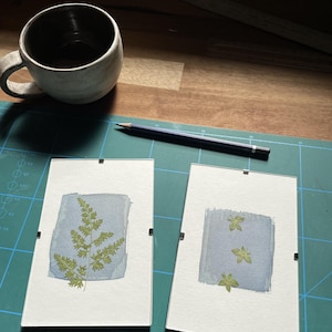 May include: Two framed botanical prints featuring pressed ferns and leaves on a light blue watercolor background. A ceramic mug and a pencil are also visible on a green cutting mat.