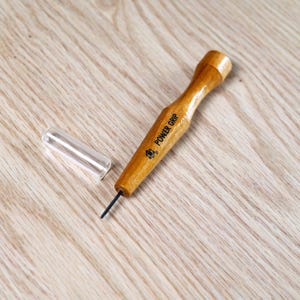 May include: A wooden awl with a clear plastic protective cap. The awl has a light brown handle with the text "POWER GRIP" printed on it. The awl's metal tip is black. The cap is off to the side.