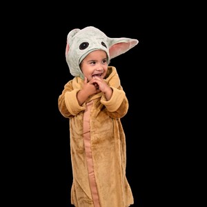 Puede incluir: Un ni&ntilde;o o ni&ntilde;a lleva una bata de felpa marr&oacute;n con una capucha de personaje verde y rosa. El ni&ntilde;o o ni&ntilde;a sonr&iacute;e y mira hacia la derecha.