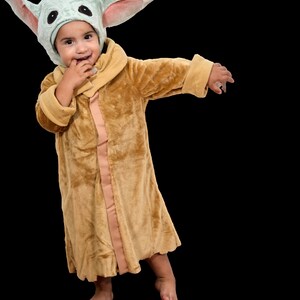 Puede incluir: Un ni&ntilde;o con una bata de felpa marr&oacute;n con un disfraz de personaje con capucha verde y rosa. El ni&ntilde;o sonr&iacute;e y se lleva la mano a la boca.