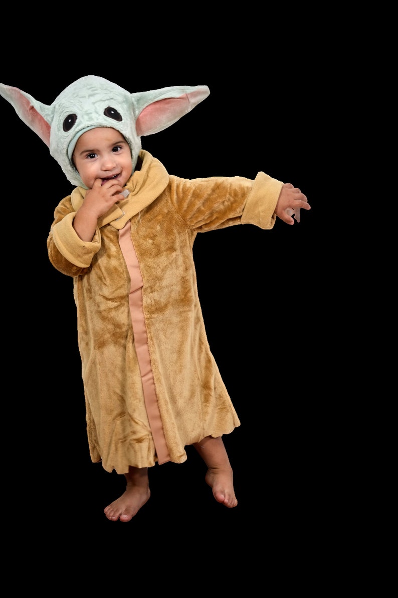 Puede incluir: Un ni&ntilde;o con una bata de felpa marr&oacute;n con un disfraz de personaje con capucha verde y rosa. El ni&ntilde;o sonr&iacute;e y se lleva la mano a la boca.