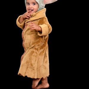 Puede incluir: Un ni&ntilde;o o ni&ntilde;a lleva una bata de color marr&oacute;n peluda y un disfraz con capucha verde y rosa con orejas grandes. El disfraz est&aacute; inspirado en el personaje Baby Yoda de la serie Star Wars.