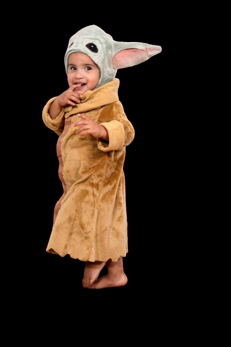Puede incluir: Un ni&ntilde;o o ni&ntilde;a lleva una bata de color marr&oacute;n peluda y un disfraz con capucha verde y rosa con orejas grandes. El disfraz est&aacute; inspirado en el personaje Baby Yoda de la serie Star Wars.