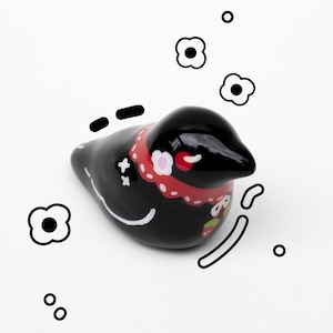 May include: A black ceramic bird ornament with a glossy finish. The bird has a red and white floral collar and white details. Black and white floral and dot decorations surround the bird on a white background.