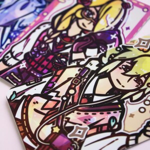 Può includere: Quattro carte illustrate colorate con personaggi in stile anime con accenti rosa acceso, viola e giallo. I personaggi indossano abiti colorati e hanno acconciature uniche.