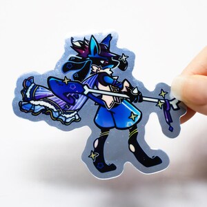 Puede incluir: Una pegatina hologr&aacute;fica con un personaje de dibujos animados azul y morado con un bast&oacute;n en forma de estrella. El personaje lleva un atuendo azul y blanco y un sombrero con una estrella en la parte superior.