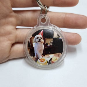 May include: A white dog wearing a red party hat sits in front of a birthday cake. The dog is in a clear plastic keychain with a silver ring.
