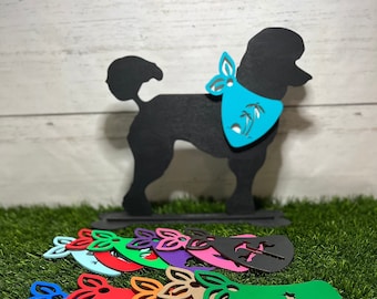 Peut inclure: Une silhouette noire de caniche portant un bandana turquoise avec des palmiers imprimés dessus. Le caniche tient une rangée de bandanas pour chien en bois colorés avec différents motifs, notamment des palmiers, des fleurs et une empreinte de patte.