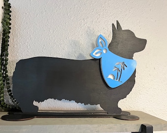 May include: A black wooden corgi dog silhouette with a light blue bandana featuring a beach scene with palm trees and a sun. The dog is standing on a base, suitable for display.
