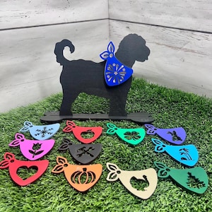 Può includere: Una silhouette nera di un cane che indossa un bandana blu con un disegno di fuochi d'artificio. Il cane è in piedi su una base di legno con 12 bandana colorati con diversi disegni, tra cui un fiocco di neve, una mela, un trifoglio, un coniglio, una palma, un cuore, una zucca, un tacchino e un albero di Natale.