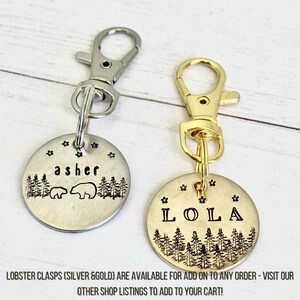 May include: Two round metal pet tags with lobster clasps. One tag is silver with the name "asher" and a bear and forest design. The other tag is gold with the name "LOLA" and a forest design. Both tags have star accents.