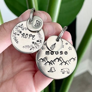May include: Two silver metal keychains. One keychain has the word "HOPE" surrounded by floral designs and a small butterfly charm. The other keychain has the word "moose" above a mountain range, a tent, and a bear.