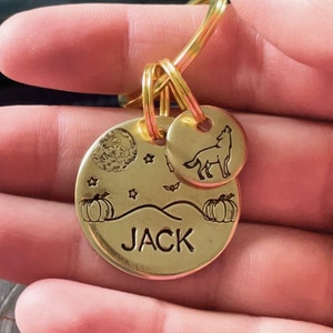 May include: A gold-colored metal keychain with two circular pendants. The larger pendant features a moon, stars, pumpkins, and the name "JACK." The smaller pendant has a howling wolf. The keychain is attached to a gold ring.
