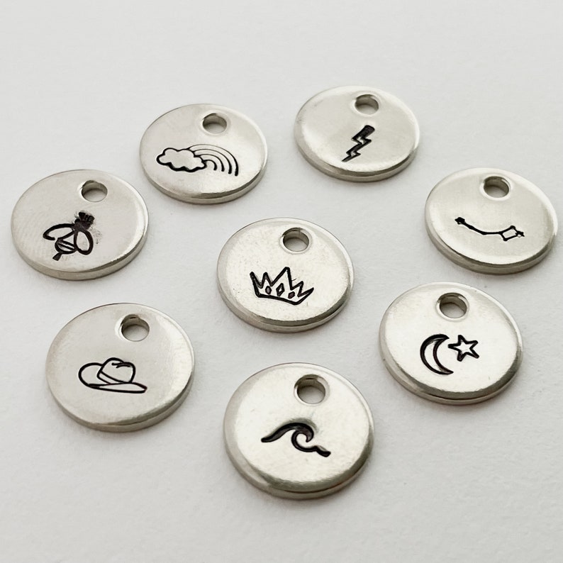 May include: A collection of small, silver-colored, round charms with various black etched designs. The charms feature images of a rainbow, lightning bolt, constellation, crown, moon and star, wave, cowboy hat, and bow and arrow.