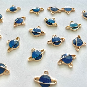 Puede incluir: Una colección de pequeños dijes dorados con forma de planeta y acabado en esmalte azul oscuro. Cada dije presenta un planeta con un anillo alrededor y un pequeño lazo para sujetarlo a joyas u otras manualidades. Los dijes están dispersos sobre una superficie blanca.