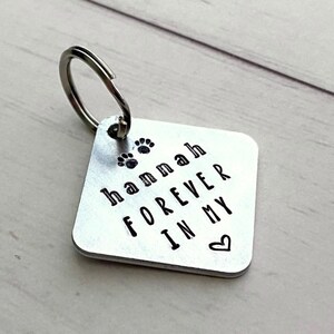 Może przedstawiać: Srebrny metalowy brelok do kluczy z kwadratową zawieszką. Zawieszka ma imię "Hannah" i frazę "FOREVER IN MY" wytłoczone na czarno, wraz z odciskami łap i symbolem serca.
