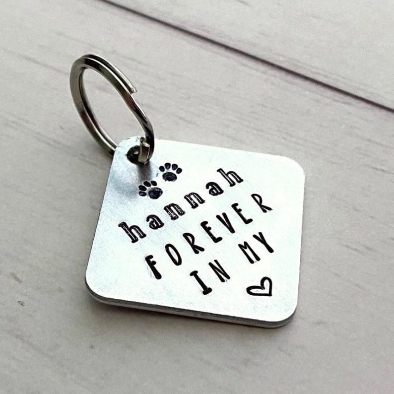 Może przedstawiać: Srebrny metalowy brelok do kluczy z kwadratową zawieszką. Zawieszka ma imię "Hannah" i frazę "FOREVER IN MY" wytłoczone na czarno, wraz z odciskami łap i symbolem serca.