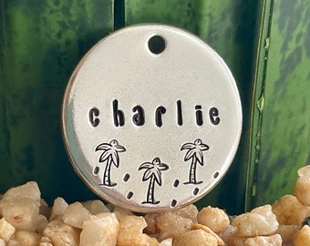 May include: A round, silver pet tag with the name "charlie" stamped in black letters. Below the name are three small palm tree designs. The tag has a hole at the top for attachment.