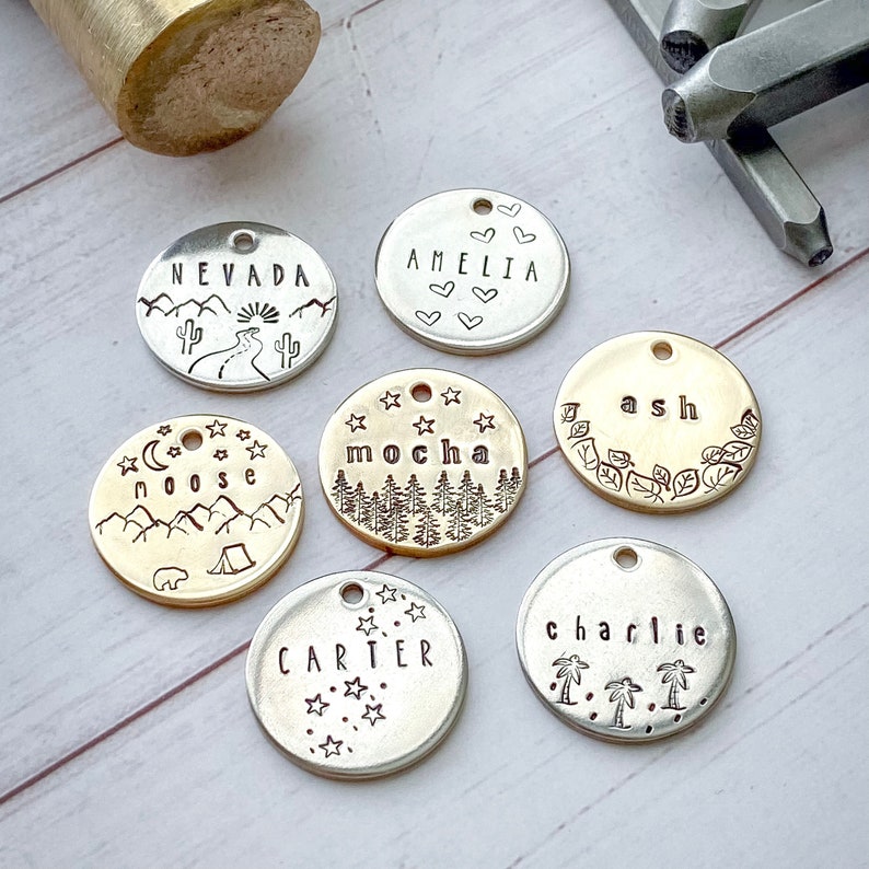 May include: A collection of stamped metal pendants with various designs and text. The pendants include names like "Amelia" and "Charlie", along with nature-themed illustrations such as mountains, trees, and cacti. A hammer and metal stamps are visible.