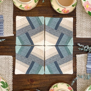 May include: Four quilted fabric coasters in a geometric pattern of beige, gray, and teal. The coasters are arranged in a square shape on a wooden table with four teacups and saucers with floral designs.
