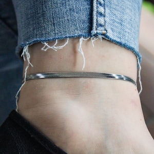 Könnte beinhalten: Ein silberfarbenes, flaches Fischgrät-Kettenarmband. Das Armband wird am Knöchel getragen, mit einer Jeans im Hintergrund. Das Armband ist ein schlichtes, elegantes Schmuckstück.