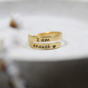 Könnte beinhalten: Goldfarbener Ring mit den Worten "Ich bin genug" und einem kleinen Herz, das in die Band graviert ist.