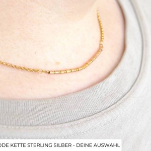 Op de afbeelding: Een gouden ketting met een rij kleine gouden kralen. De ketting wordt gedragen door een persoon met een lichtbruin shirt.