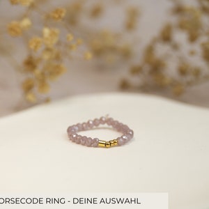 Könnte beinhalten: Ein zarter Ring aus kleinen, hellvioletten Perlen mit zwei goldenen Abstandshaltern. Der Ring liegt auf einer weißen Oberfläche.