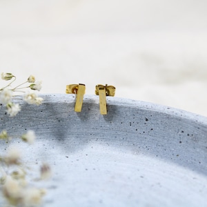 Könnte beinhalten: Ein Paar goldene Barren-Ohrstecker auf einer hellgrauen, strukturierten Oberfläche.