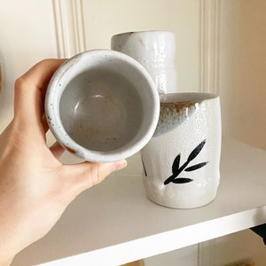 May include: Three white ceramic mugs with black leaf designs. The mugs are on a white shelf.