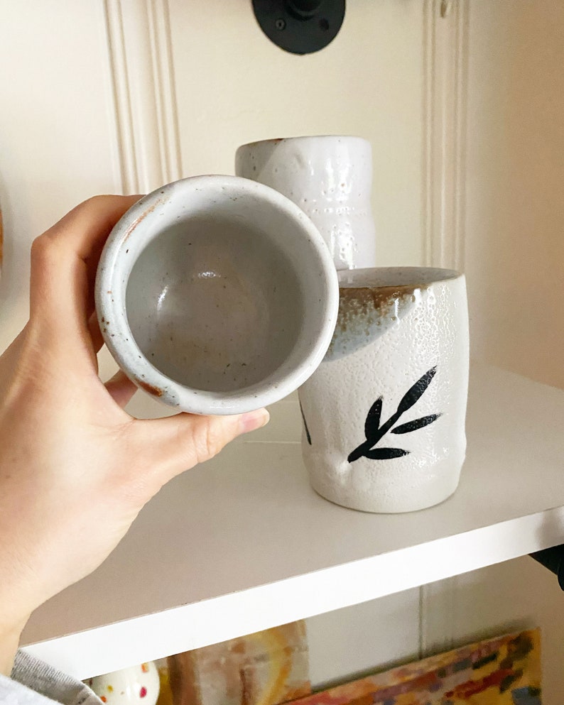 May include: Three white ceramic mugs with black leaf designs. The mugs are on a white shelf.