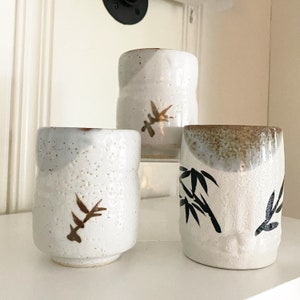 May include: Three white ceramic teacups with different designs. The first cup has a brown leaf design, the second cup has a brown leaf design, and the third cup has a black bamboo design.