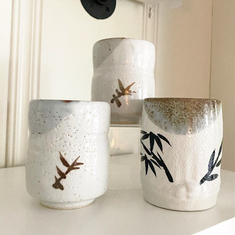 May include: Three white ceramic teacups with different designs. The first cup has a brown leaf design, the second cup has a brown leaf design, and the third cup has a black bamboo design.