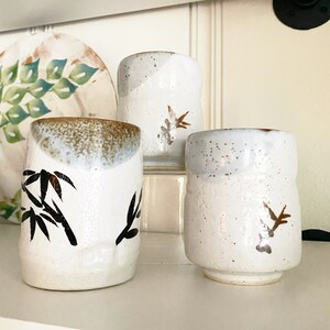 May include: Three white ceramic mugs with brown and black designs. The mugs have a speckled glaze and are decorated with stylized bamboo and floral patterns.