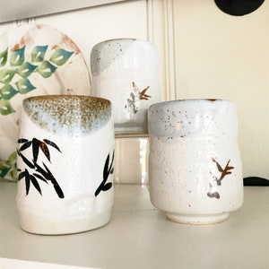 May include: Three white ceramic mugs with brown and black designs. The mugs have a speckled glaze and are decorated with bamboo and bird motifs.