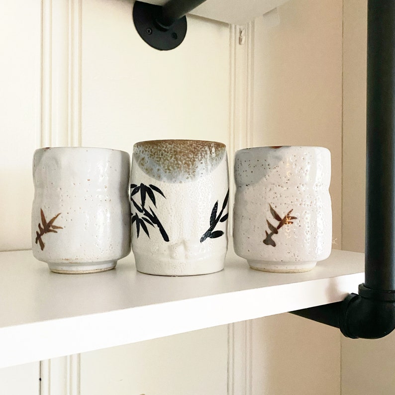 May include: Three white ceramic teacups with black and brown designs. The cups are sitting on a white shelf.