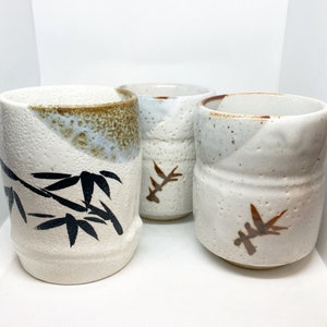 May include: Three white ceramic teacups with brown and black designs. The cups have a speckled glaze and are decorated with stylized bamboo leaves.