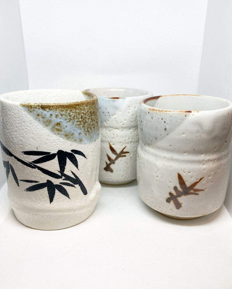 May include: Three white ceramic teacups with brown and black designs. The cups have a speckled glaze and are decorated with stylized bamboo leaves.