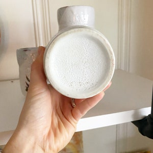 May include: White ceramic vase with a speckled glaze. The vase has a round bottom and a slightly flared opening.