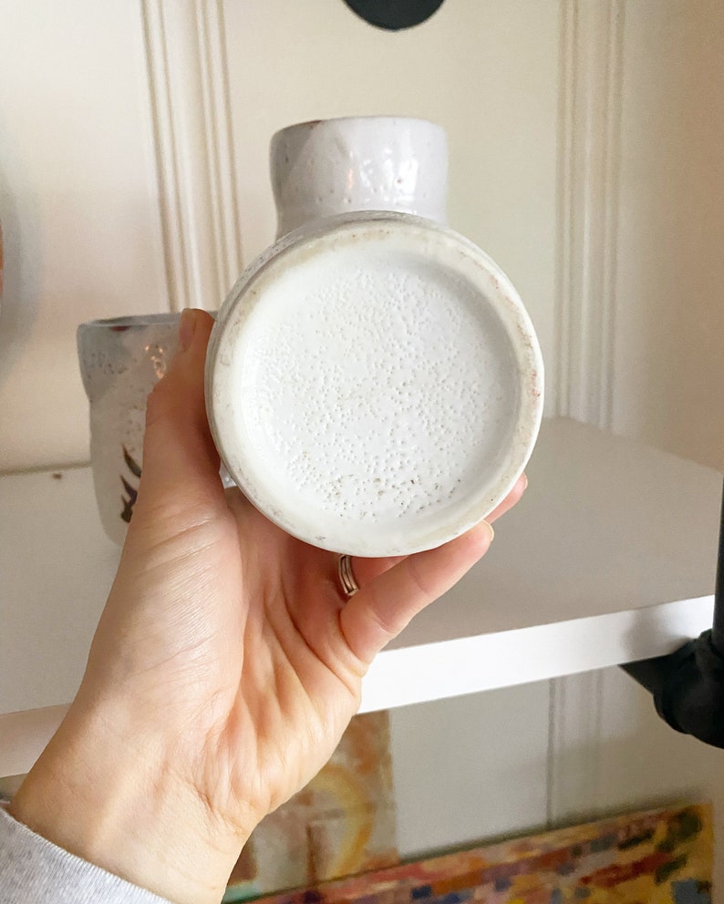 May include: White ceramic vase with a speckled glaze. The vase has a round bottom and a slightly flared opening.