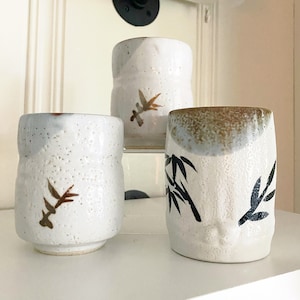 May include: Three white ceramic mugs with brown and black painted designs. The mugs have a textured surface and are decorated with stylized plant motifs.