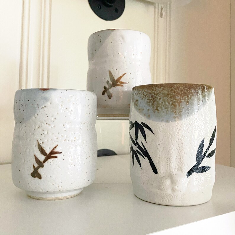 May include: Three white ceramic mugs with brown and black painted designs. The mugs have a textured surface and are decorated with stylized plant motifs.