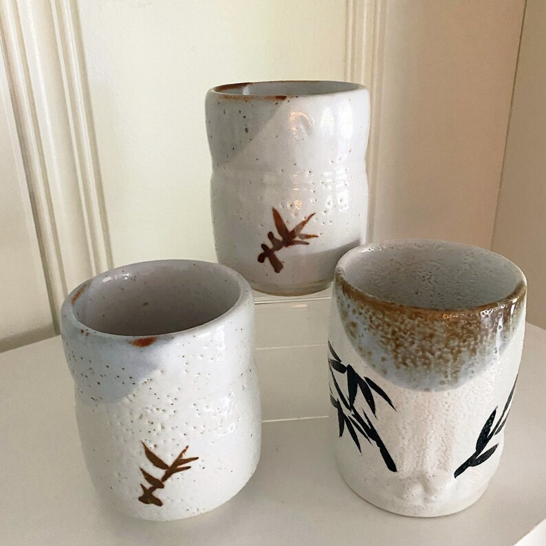 May include: Three white ceramic cups with brown and black painted designs. The cups have a speckled glaze and are slightly rounded.