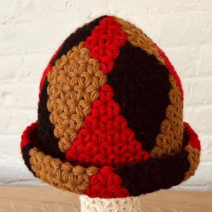 Puede incluir: Un sombrero de ganchillo hecho a mano con un patrón geométrico en rojo, negro y marrón. El sombrero tiene un borde enrollado y se exhibe sobre un soporte crema con un patrón de estrellas. Un accesorio único y colorido.