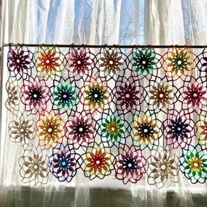 Könnte beinhalten: Ein farbenfroher gehäkelter Gardinen-Volant mit einem Regenbogen an Farben, darunter Rot, Orange, Gelb, Grün, Blau, Indigo und Violett. Der Volant ist aus einem zarten, spitzigen Stoff gefertigt und hängt an einer weißen Stange.